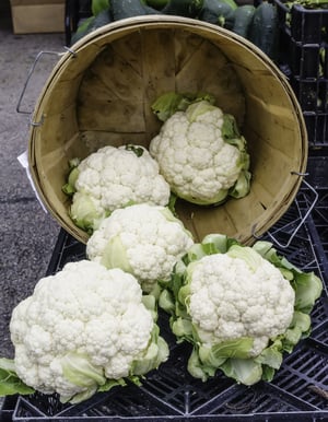 Five white cauliflowers, two still in the basket, on display cornucopia-style at a farmers market (foreground focus)
