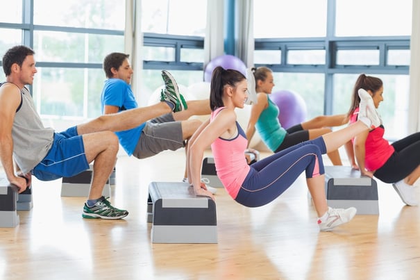 Full length side view of instructor with fitness class performing step aerobics exercise in gym.jpeg